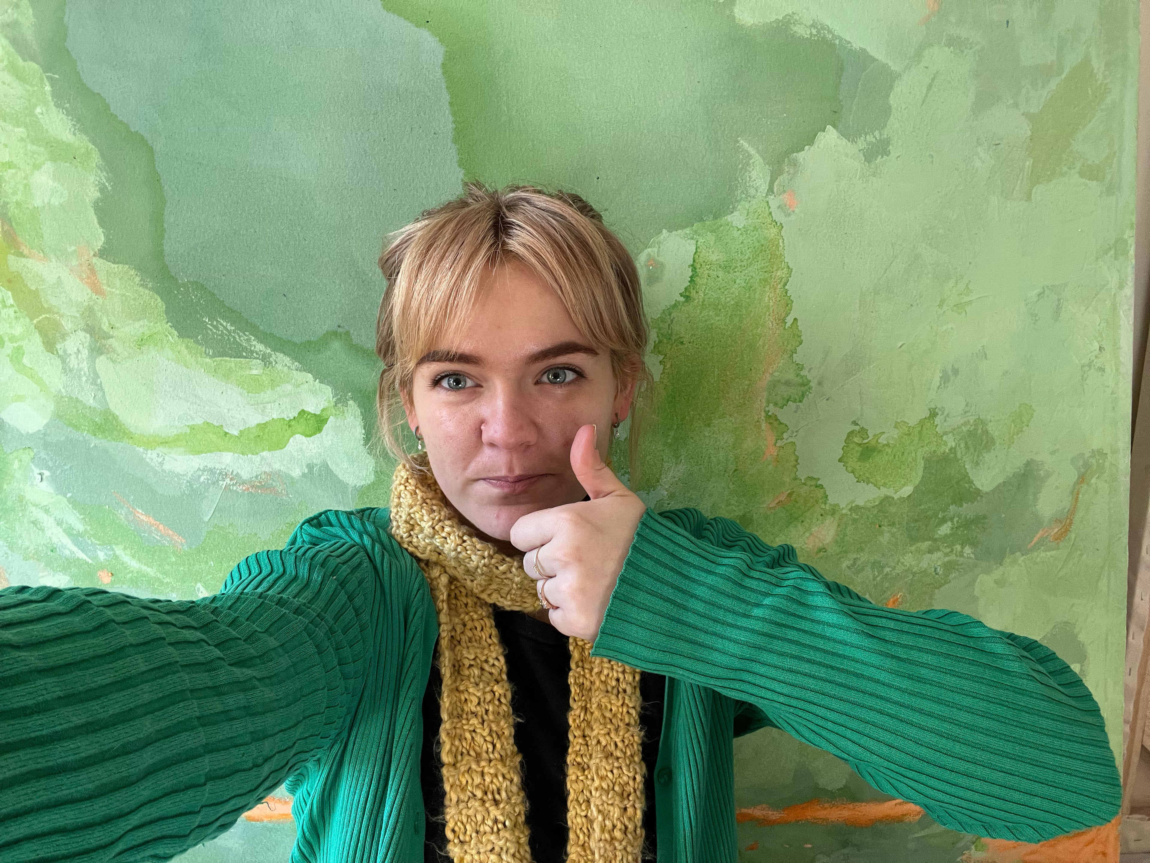Person wearing a green sweater and yellow scarf in front of a green abstract painting